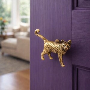 Könnte beinhalten: Ein messingfarbener Türgriff in Katzenform ist an einer violetten Tür angebracht. Der Griff ist eine detaillierte Skulptur einer Katze mit eingerolltem Schwanz. Die Tür ist einfarbig, und der Griff sorgt für eine dekorative Note.