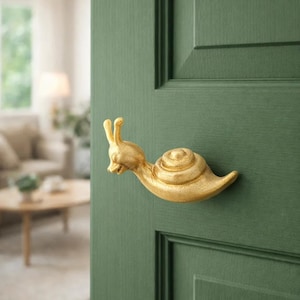 May include: A whimsical gold-colored snail door handle mounted on a green door. The snail's body is detailed with a spiral shell and expressive face. The door is painted a deep green, with a glimpse of a living room in the background.