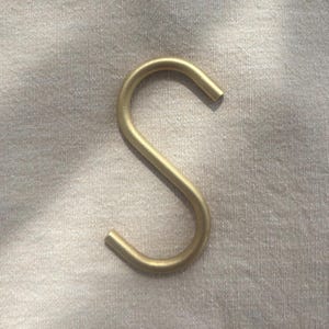 May include: A gold-colored metal S-hook, curved at both ends. The hook is made of a solid, smooth material and is set against a neutral-toned fabric background. The hook is designed for hanging items.