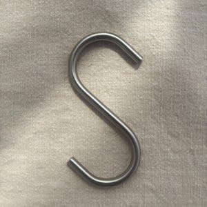 Op de afbeelding: Een enkele, glanzende, zilverkleurige S-haak. De haak is gemaakt van metaal en heeft een glad, gebogen ontwerp. Het wordt waarschijnlijk gebruikt om items op te hangen. De achtergrond is een neutrale, getextureerde stof.