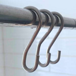 May include: Three shiny, silver-colored S-hooks hanging from a metal rod. The hooks are curved and have a simple, functional design. The background is a light, blurred white.