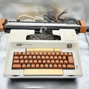 May include: A vintage Smith-Corona Coronamatic typewriter. The typewriter has a cream-coloured body with brown keys and accents. The keys are arranged in a QWERTY layout. The typewriter has a carriage return lever and a paper feed roller.