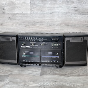 May include: A black Philips boombox featuring dual cassette decks, integrated speakers, and a radio tuner. The unit has a silver and black design with numerous dials and buttons. The "Philips" brand name is displayed prominently on the front.