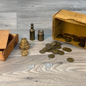 Op de afbeelding: Een assortiment antieke messing gewichten, munten en een kleine houten kist met een deksel. De gewichten zijn in verschillende vormen en maten, waaronder een stapel afgestudeerde gewichten. Een open kartonnen doos onthult meer munten.