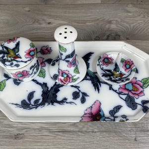 Op de afbeelding: Een vintage porseleinen toilettafelset met een rechthoekige lade, een kleine pot met deksel, een zoutstrooier en een klein schaaltje. De set is versierd met een bloemen- en vogelontwerp in roze, blauw, groen en goud op een witte achtergrond.