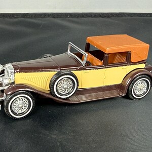 May include: A vintage-style toy car in brown, yellow, and orange. The car features white-walled tires, a brown convertible top, and chrome detailing. The car's body is a combination of brown and yellow, with a detailed front grille.