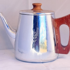 Peut inclure: Une théière en métal argenté poli avec un corps conique orné d'un motif gravé en forme d'épi de blé. La théière est dotée d'un bouton de couvercle en bois et d'une grande poignée angulaire en bois fixée sur le côté, avec un bec verseur.