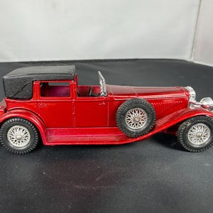 May include: A red vintage car model with a black convertible top. The car has silver-colored wheels and a spare tire. The model car is a classic design, with detailed features and a glossy finish.