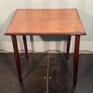 Peut inclure: Une table d'appoint rectangulaire dotée d'un plateau en bois clair au grain apparent, entouré d'une bordure en bois plus foncé. La table repose sur quatre pieds en bois foncé, fins et fuselés, s'étendant depuis les coins du cadre.