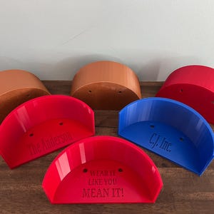 May include: A collection of semi-circular, brim-shaped items in red, blue, and brown. Some have text: "The Anderson", "C.J. Inc.", and "WEAR IT LIKE YOU MEAN IT!". They are displayed on a dark wooden surface.