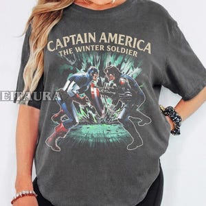 May include: Dark gray t-shirt featuring a graphic of Captain America and the Winter Soldier in a battle scene. The text "CAPTAIN AMERICA THE WINTER SOLDIER" is printed across the top in a vintage style.