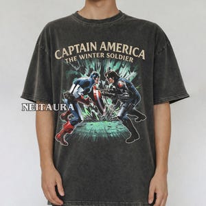 May include: Dark grey t-shirt with a graphic of Captain America and the Winter Soldier in combat. The text "CAPTAIN AMERICA THE WINTER SOLDIER" is printed above the image. The shirt has short sleeves and a crew neck.