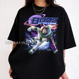 May include: Black t-shirt with a graphic of Buzz Lightyear in a dynamic pose, with the words "Buzz Lightyear" in a stylized font. The design includes a planet and space-themed elements, with a vibrant colour palette of green, purple, and white.