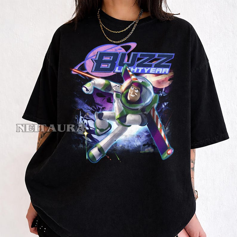 May include: Black t-shirt with a graphic of Buzz Lightyear in a dynamic pose, with the words "Buzz Lightyear" in a stylized font. The design includes a planet and space-themed elements, with a vibrant colour palette of green, purple, and white.