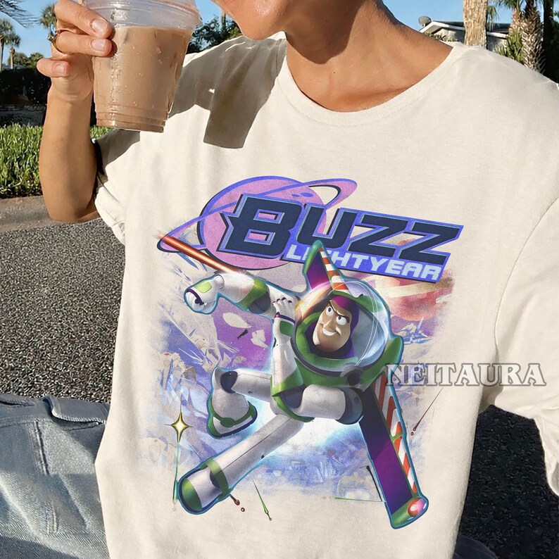 May include: Cream-coloured t-shirt with a graphic of Buzz Lightyear from Toy Story. The design features the words "BUZZ LIGHTYEAR" in a purple and blue font, with Buzz in his green and white spacesuit. The shirt has a crew neck.
