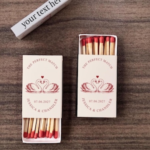 May include: Two rectangular matchboxes with cream-colored covers featuring red ink illustrations of two swans forming a heart shape. The text reads THE PERFECT MATCH, JESSICA & CHANDLER, and 07.06.2027. The matches have red tips. One box is open, showing the matches inside.