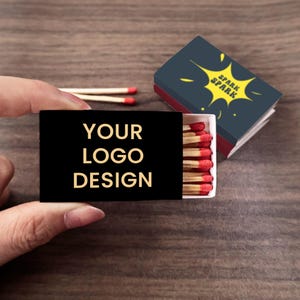 May include: A black matchbox with the text "YOUR LOGO DESIGN" in gold lettering. The matchbox is open, revealing red-tipped matches. A second matchbox with a yellow starburst design and the word "SPARK" is visible.