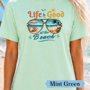 Pu&ograve; includere: T-shirt verde menta con la scritta "Life is Good at the Beach". La grafica presenta occhiali da sole con una scena di spiaggia all'interno, tra cui una sedia a sdraio, un ombrellone e palme. La maglietta &egrave; di colore pastello chiaro.