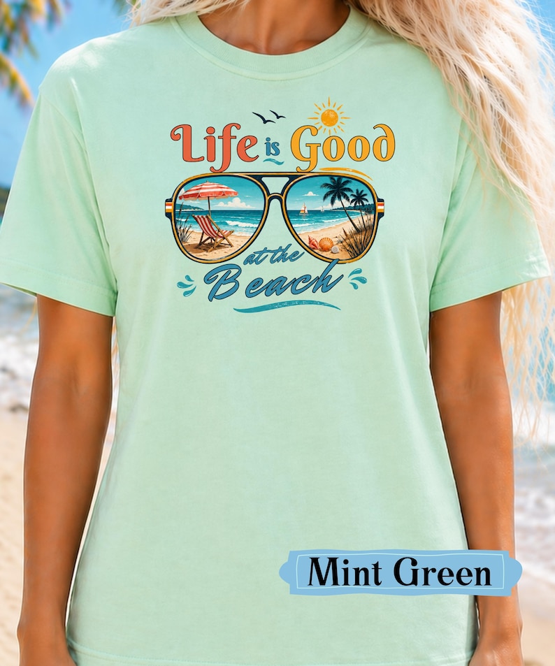 Pu&ograve; includere: T-shirt verde menta con la scritta "Life is Good at the Beach". La grafica presenta occhiali da sole con una scena di spiaggia all'interno, tra cui una sedia a sdraio, un ombrellone e palme. La maglietta &egrave; di colore pastello chiaro.