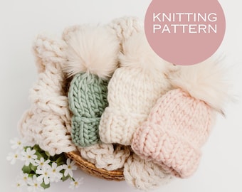 Könnte beinhalten: Drei gestrickte Mützen in einem Korb vor weißem Hintergrund. Die Mützen sind grün, weiß und rosa. Jede Mütze hat einen flauschigen weißen Bommel oben. Der Korb ist aus geflochtenem Weidengeflecht.