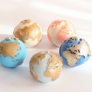 Peut inclure: Cinq objets décoratifs sphériques ressemblant à des globes miniatures. Les globes présentent des continents dorés sur des fonds bleus, blancs et roses. Les objets sont disposés sur une surface blanche.
