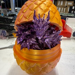 Puede incluir: Un huevo de dragón naranja impreso en 3D con una superficie texturizada y escamosa que está parcialmente abierto. Dentro del huevo hay una figura de dragón articulada de color morado metálico con púas y cuernos detallados.