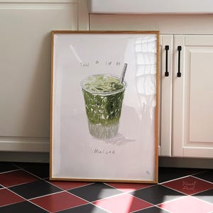 Può includere: Una stampa incorniciata di una bevanda al matcha in una tazza trasparente con una cannuccia. Il testo "TAKE A SIP OF MATCHA" è sopra e sotto la bevanda. La stampa è in una cucina con mobili bianchi e un pavimento a scacchi rosso e nero.