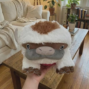 Puede incluir: Un peluche con forma de bisonte blanco y marrón, con ojos grandes y expresivos. El juguete tiene una textura suave y presenta pezuñas marrones y una cara gris. El bisonte es un personaje de la serie animada Avatar: La leyenda de Aang.