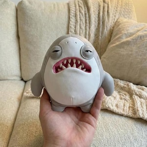 Op de afbeelding: Een pluche haaienspeelgoed met een humeurige uitdrukking. De haai is voornamelijk grijs en wit, met een roze binnenkant van de mond en scherpe witte tanden. Het speelgoed heeft kleine vinnen en wordt in een hand gehouden.