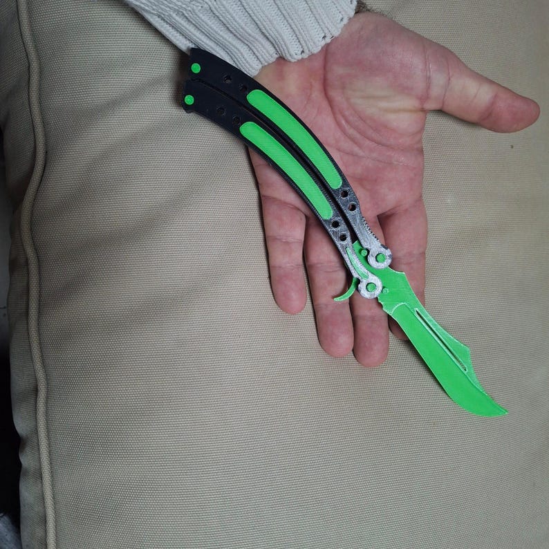 May include: A butterfly knife with a black handle and bright green accents. The blade is also bright green. The knife is open and ready for use. The knife is held in a hand.