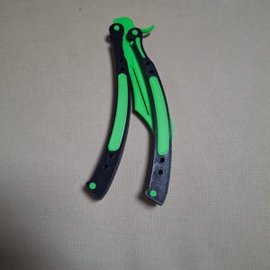 May include: A butterfly knife with a black handle and a bright green blade. The handle has a curved design with a series of small holes. The blade is also green and has a curved shape. The knife is lying on a neutral background.