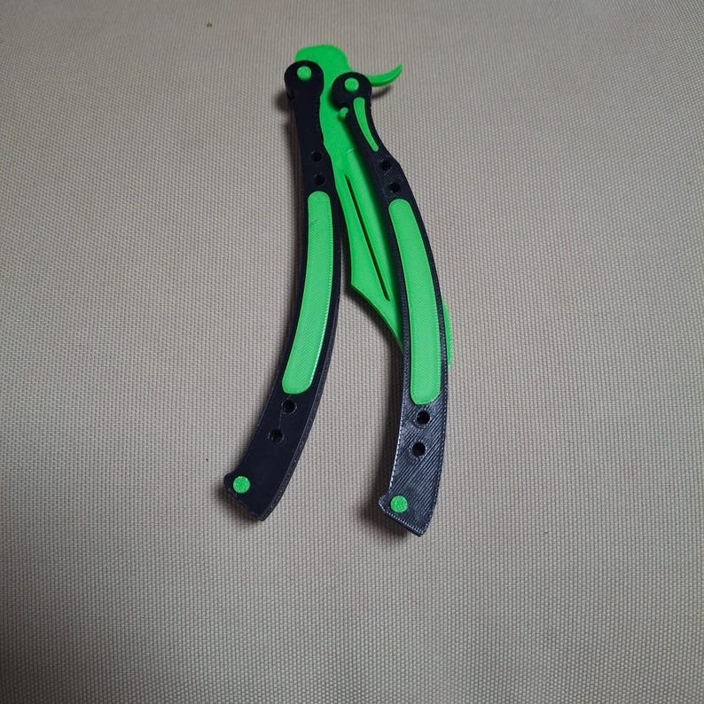 May include: A butterfly knife with a black handle and a bright green blade. The handle has a curved design with a series of small holes. The blade is also green and has a curved shape. The knife is lying on a neutral background.
