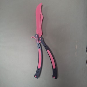 May include: A butterfly knife with a bright pink blade and a dark gray handle featuring pink accents. The handle is composed of two pivoting arms connected to the blade base, with visible circular cutouts and hardware along the frame.