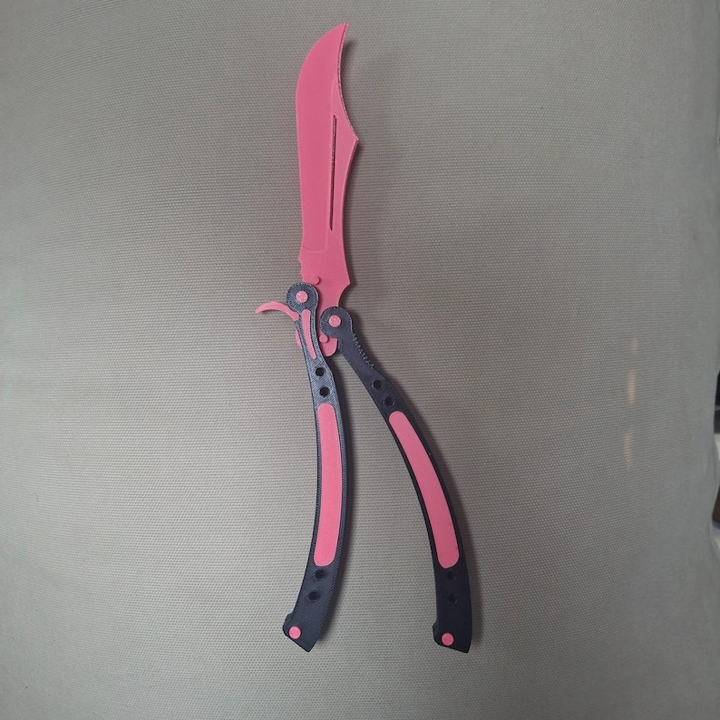 May include: A butterfly knife with a bright pink blade and a dark gray handle featuring pink accents. The handle is composed of two pivoting arms connected to the blade base, with visible circular cutouts and hardware along the frame.