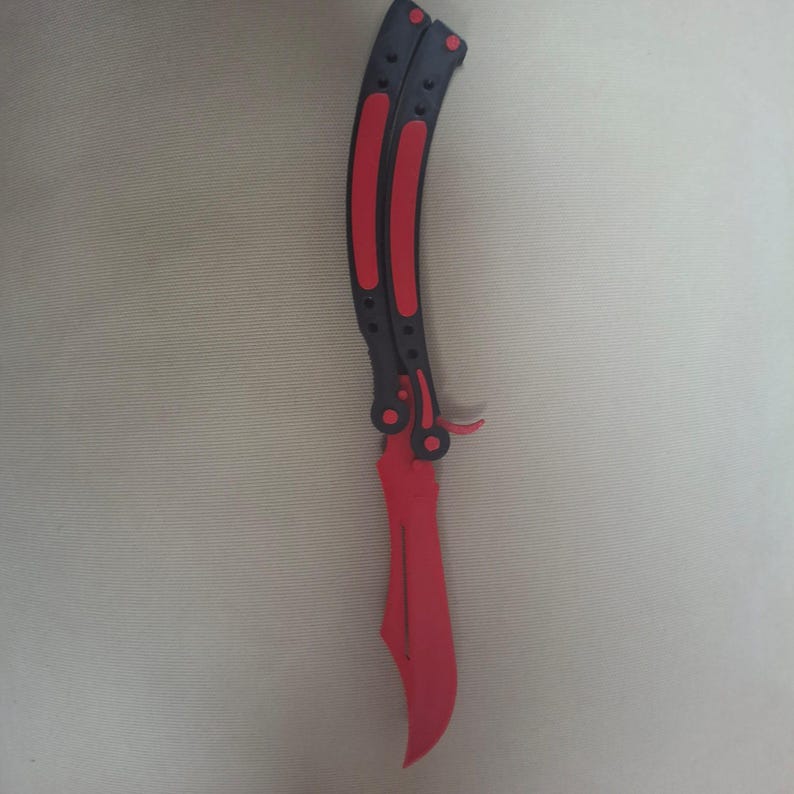 May include: A butterfly knife trainer featuring a matte black handle with vibrant red rubberized grip inserts. The blade is solid red with a curved, non-sharpened edge. The folding mechanism includes two handle segments connected by a pivot pin assembly.