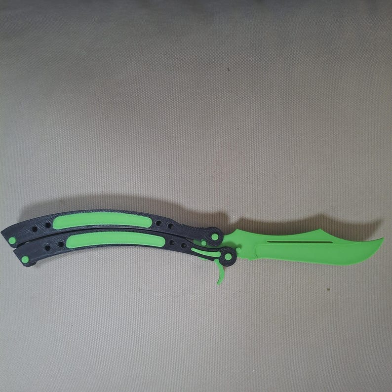 May include: A butterfly knife trainer featuring a bright green blade and a black handle with green accents. The handle consists of two pivoting sections with textured surfaces, and the blade has a curved edge and a small cutout detail.