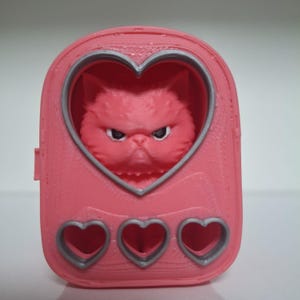 May include: A pink cat carrier with a heart-shaped window showcasing a grumpy cat. The carrier is rectangular with three heart-shaped cutouts at the base. The cat's face is detailed with furrowed brows and a stern expression, giving it a unique look.