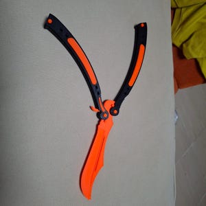 May include: A butterfly knife trainer featuring a bright orange blade and a black handle with orange accents. The folding mechanism includes two handle segments that pivot around the central orange blade, showcasing a dual-tone design.