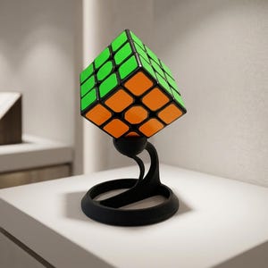 May include: A three-dimensional puzzle cube featuring bright green and orange square panels on its visible faces, balanced at a corner point on a black, curved, circular-based stand that elevates the cube above the flat surface.