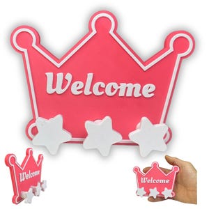 Puede incluir: Un perchero de pared con forma de corona rosa que presenta el texto blanco "Welcome" en letra cursiva. El diseño incluye tres ganchos blancos con forma de estrella unidos al borde inferior, con un borde blanco que perfila la corona.