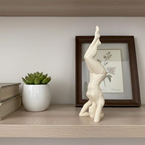 May include: A cream-colored ceramic figurine depicting a person in a headstand yoga pose. The figure has a smooth, matte finish and is shown in a vertical, inverted position with legs extended upward and arms bent at the elbows for support.