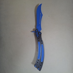 May include: A butterfly knife featuring a curved, bright blue blade with a central cutout. The handle is constructed from two black segments, each inlaid with a matching blue strip, connected by a pivot mechanism at the base of the blade.