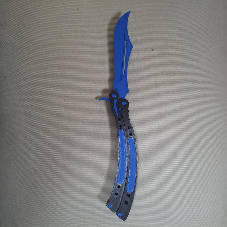 May include: A butterfly knife featuring a curved, bright blue blade with a central cutout. The handle is constructed from two black segments, each inlaid with a matching blue strip, connected by a pivot mechanism at the base of the blade.