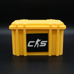 May include: A bright yellow rectangular storage case with a ribbed exterior design. Centered on the front is a black rectangular patch featuring the white text CKS with a small white silhouette of a person aiming a rifle integrated into the letter K.