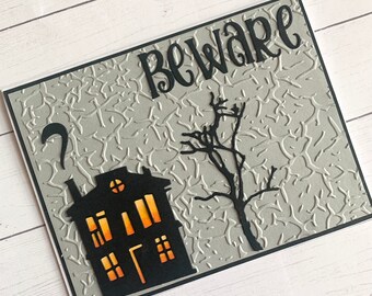May include: A gray card with a black silhouette of a house with lit windows and a bare tree. The text "Beware" is written in black at the top of the card.