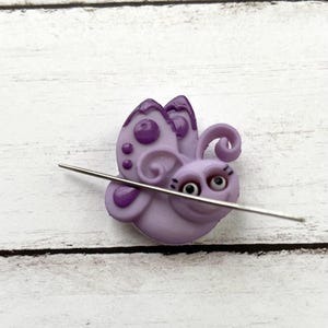 May include: Purple butterfly-shaped needle minder with a smiling face and googly eyes. The needle minder is holding a needle in its mouth.