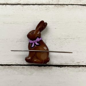 May include: A chocolate bunny with a purple bow, used as a yarn needle holder. The needle is threaded through the bunny's body.
