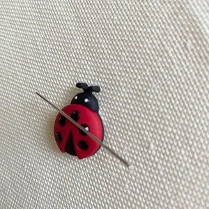 May include: A red and black ladybug-shaped needle minder with black dots and a silver needle threaded through it.