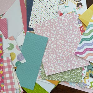 May include: A collection of patterned paper scraps in various shapes and sizes. Designs include polka dots, floral patterns, gingham, and geometric shapes. Colors include pink, blue, green, yellow, and white. Some papers feature illustrations of bicycles and flowers.