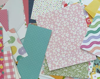May include: A collection of patterned paper scraps in various shapes and sizes. Designs include polka dots, floral patterns, gingham, and geometric shapes. Colors include pink, blue, green, yellow, and white. Some papers feature illustrations of bicycles and flowers.